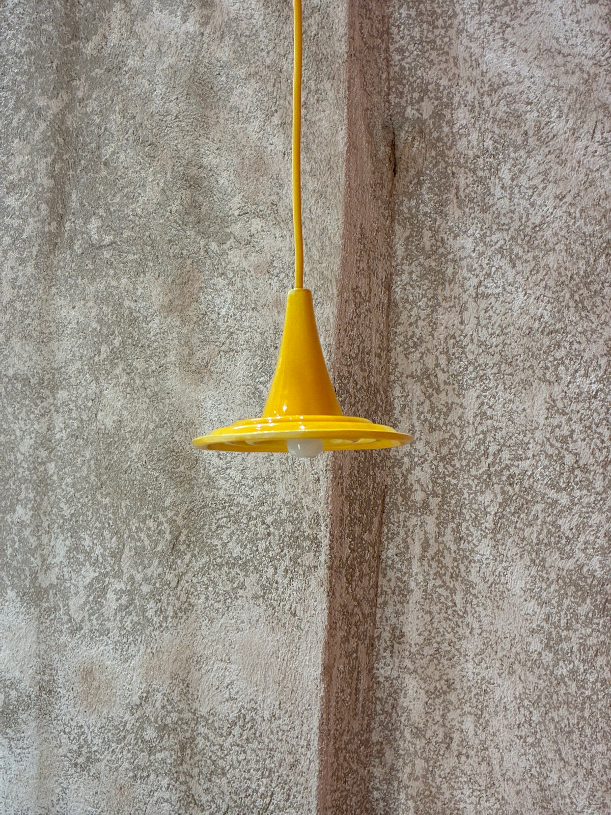 Petite suspension Ondine en céramique jaune, vue de profil devant un mur texturé gris.
