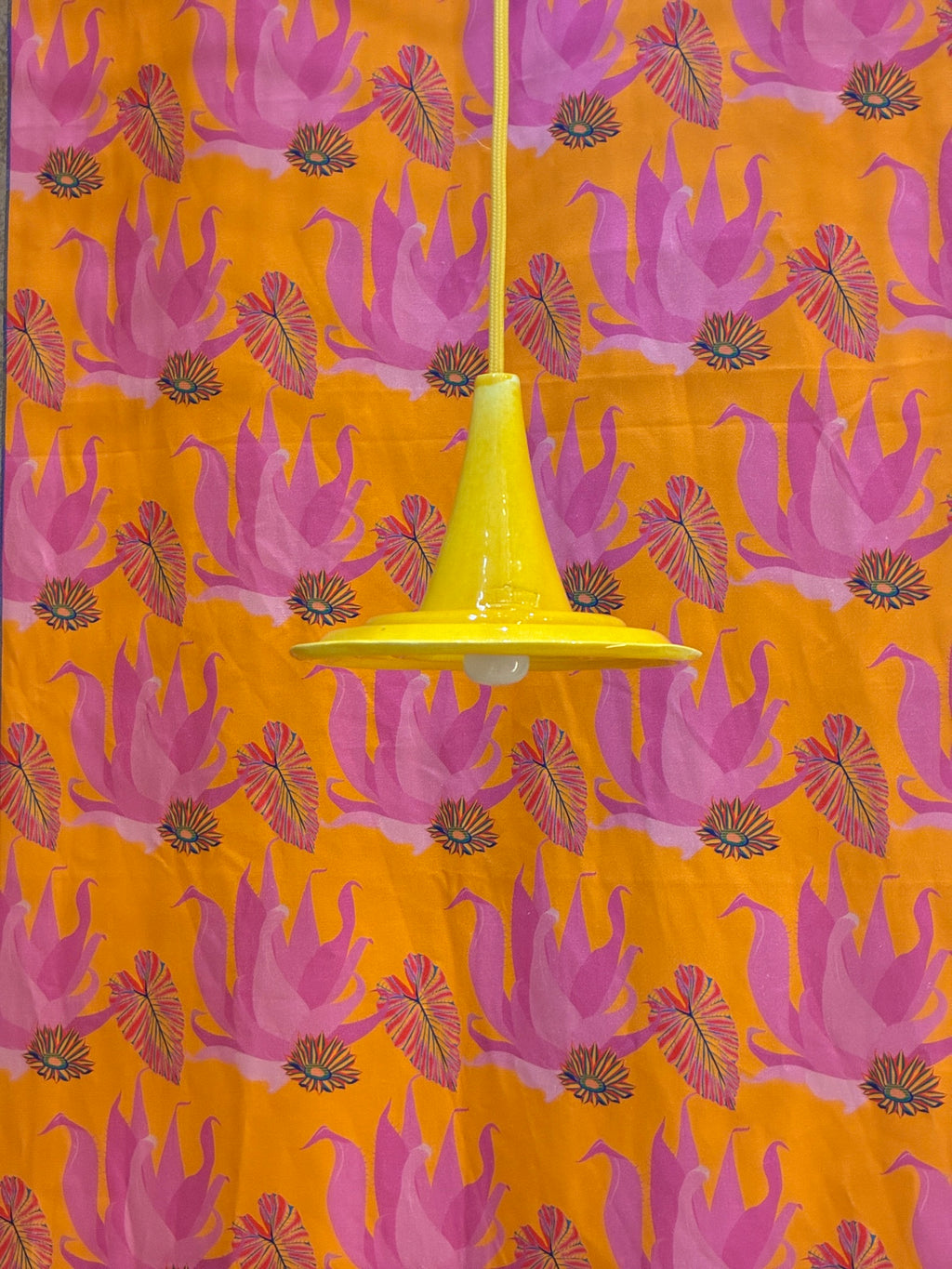 Suspension Ondine Jaune Soleil et câble jaune devant un fond à motifs roses et orange.