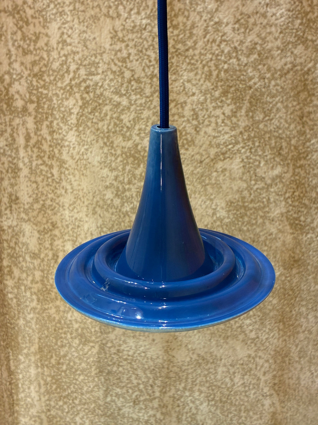 Suspension Ondine en céramique bleue avec câble bleu, vue de trois quarts devant un mur texturé. 