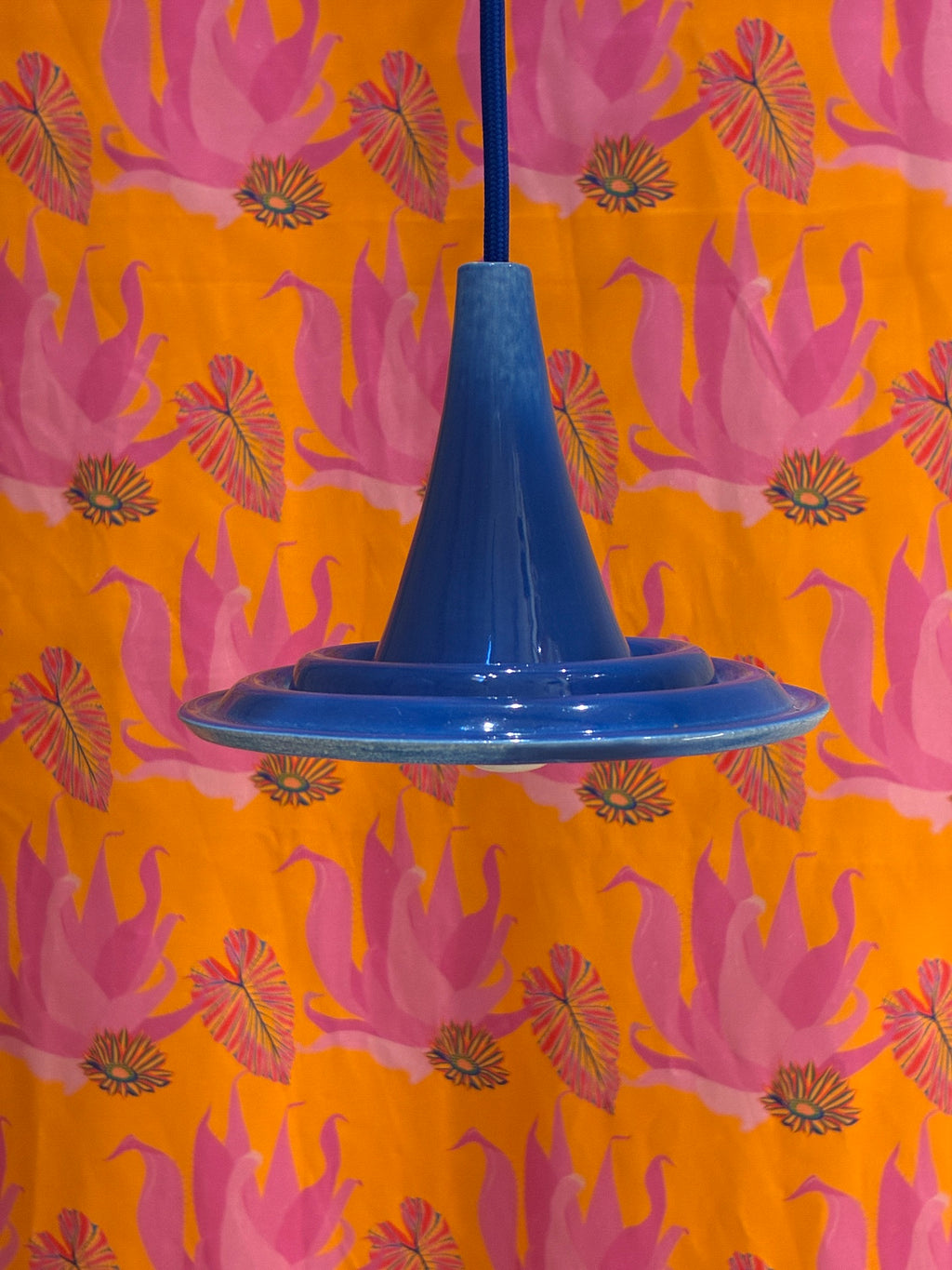  Petite suspension Ondine bleue vue de profil devant un fond à motifs roses et orange. 